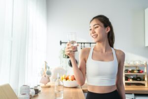 Krásná asijská žena ve sportovním oblečení pije vodu po cvičení doma.